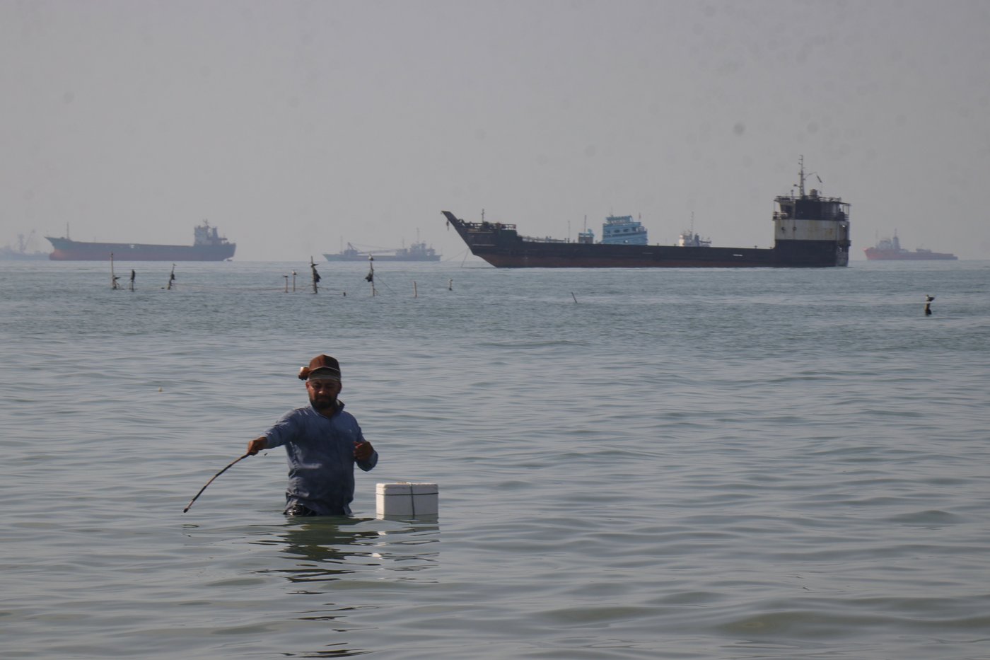 A man stands in the water, appearing to fish, as bulk carriers, cargo ships, and service vessels line the horizon in the Strait of Hormuz off Bandar Abbas, Iran, Monday, April 27, 2026.(Razieh Poudat/ISNA via AP)