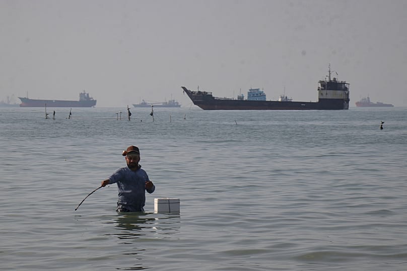 A man stands in the water as bulk carriers, cargo ships, and service vessels line the horizon in the Strait of Hormuz off Bandar Abbas, Iran, Monday, April 27, 2026 Razieh Poudat/AP