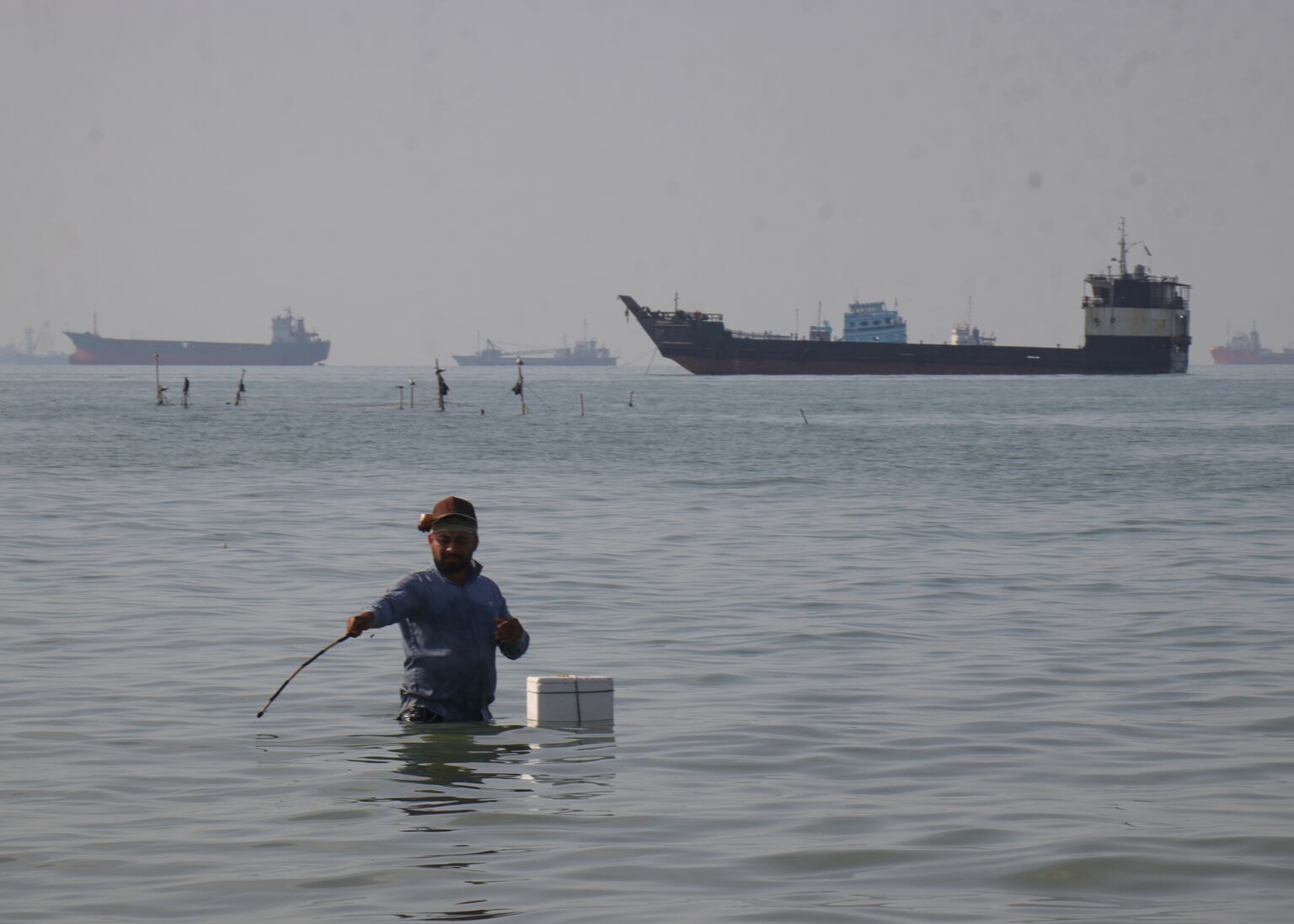 A man stands in the water, appearing to fish, as bulk carriers, cargo ships, and service vessels line the horizon in the Strait of Hormuz off Bandar Abbas, Iran (Razieh Poudat/AP)